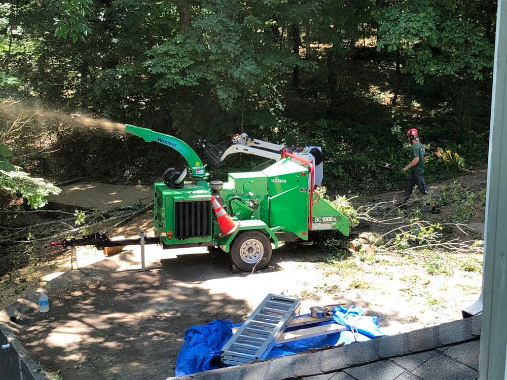 Tree chipper in use, clearing branches in wooded area, with worker and equipment visible.