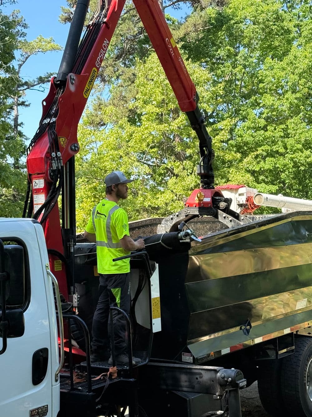 Worker operating a crane on a truck to load materials surrounded by trees.