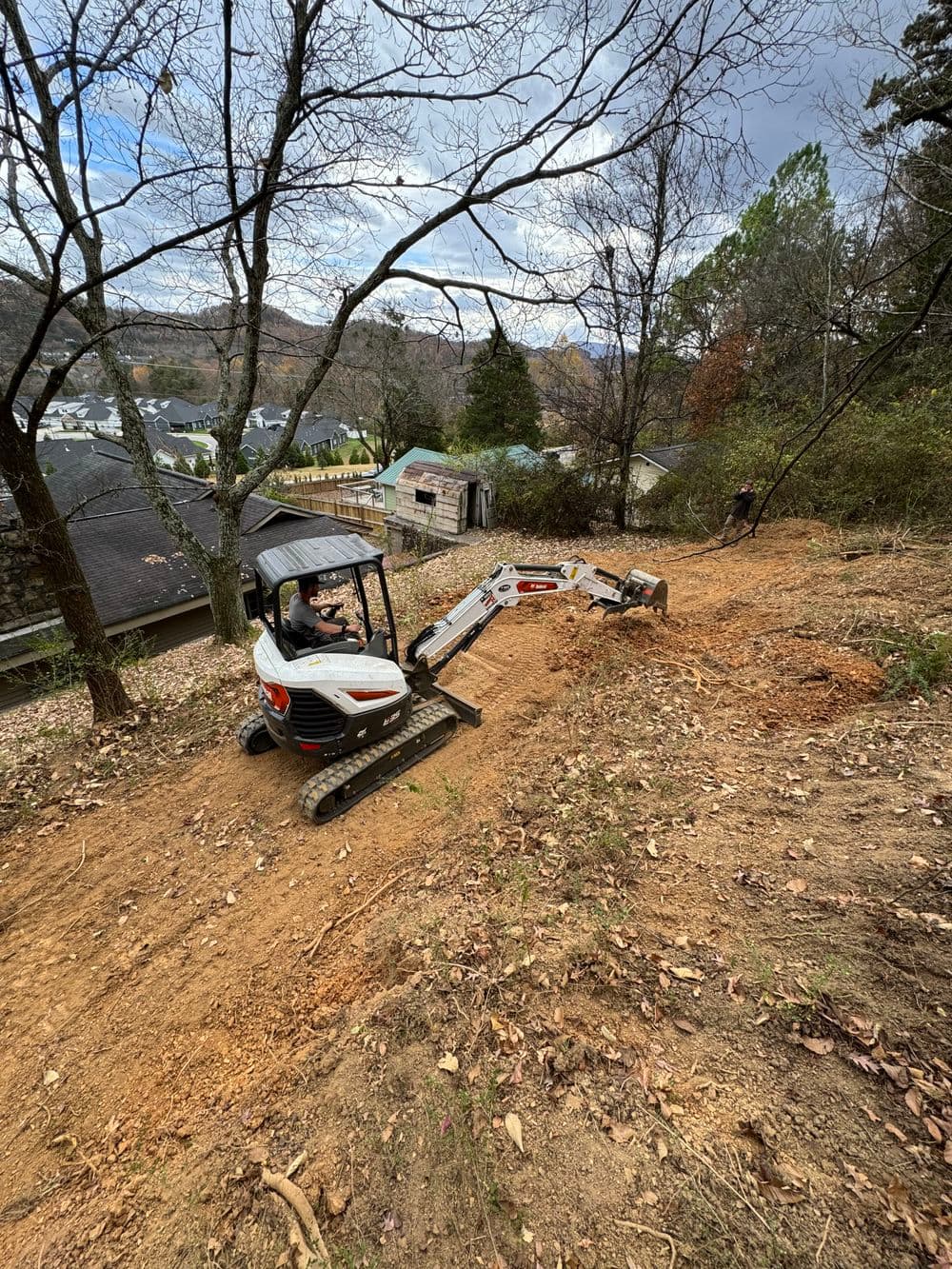 Excavator clearing land on a hillside with houses and trees in the background.