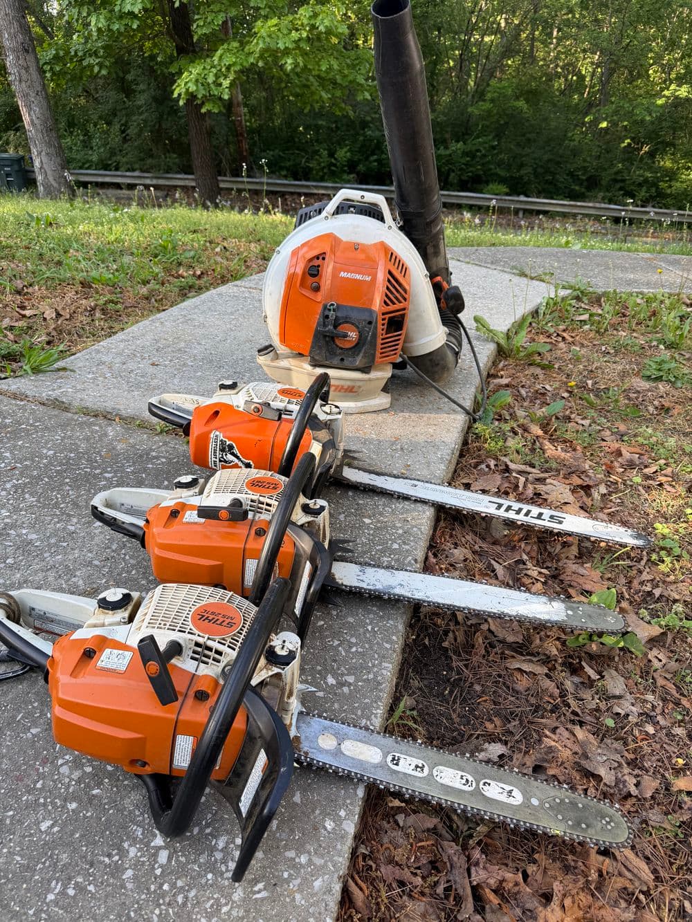 Chainsaws and leaf blower arranged on a path in a wooded area, showcasing outdoor tools.