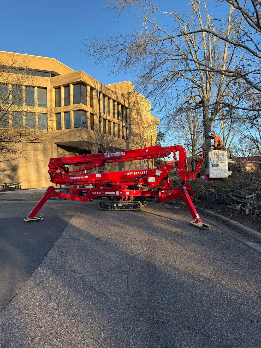 Red boom lift working near a commercial building during daylight, with a worker in safety gear.
