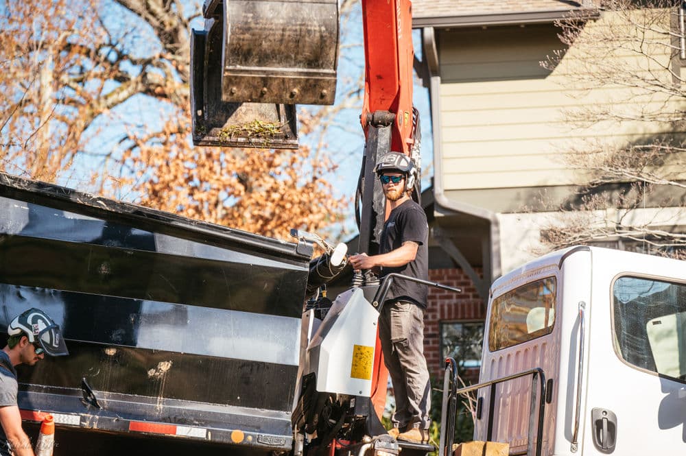 Man operating a dump truck's crane to load debris at a residential construction site.